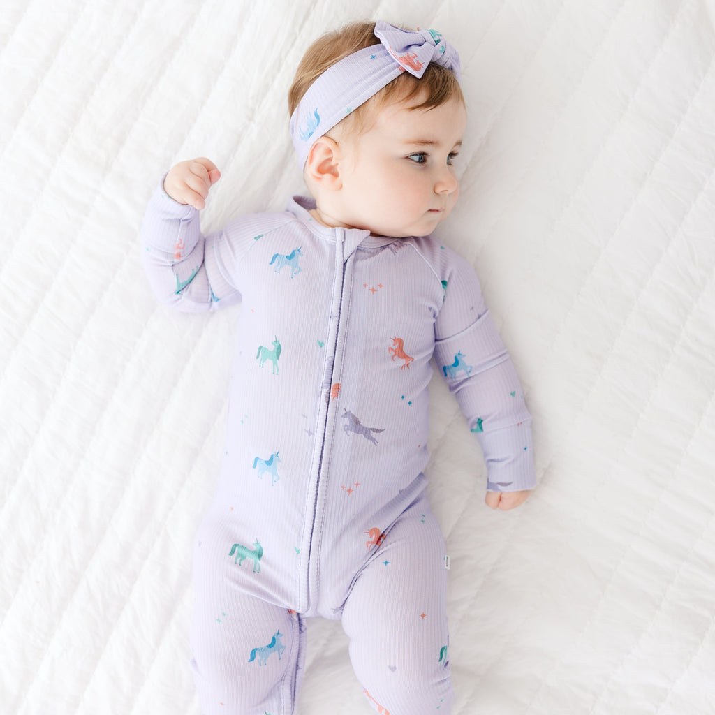 Baby wearing a light purple onesie with colorful patterns on a white textured surface