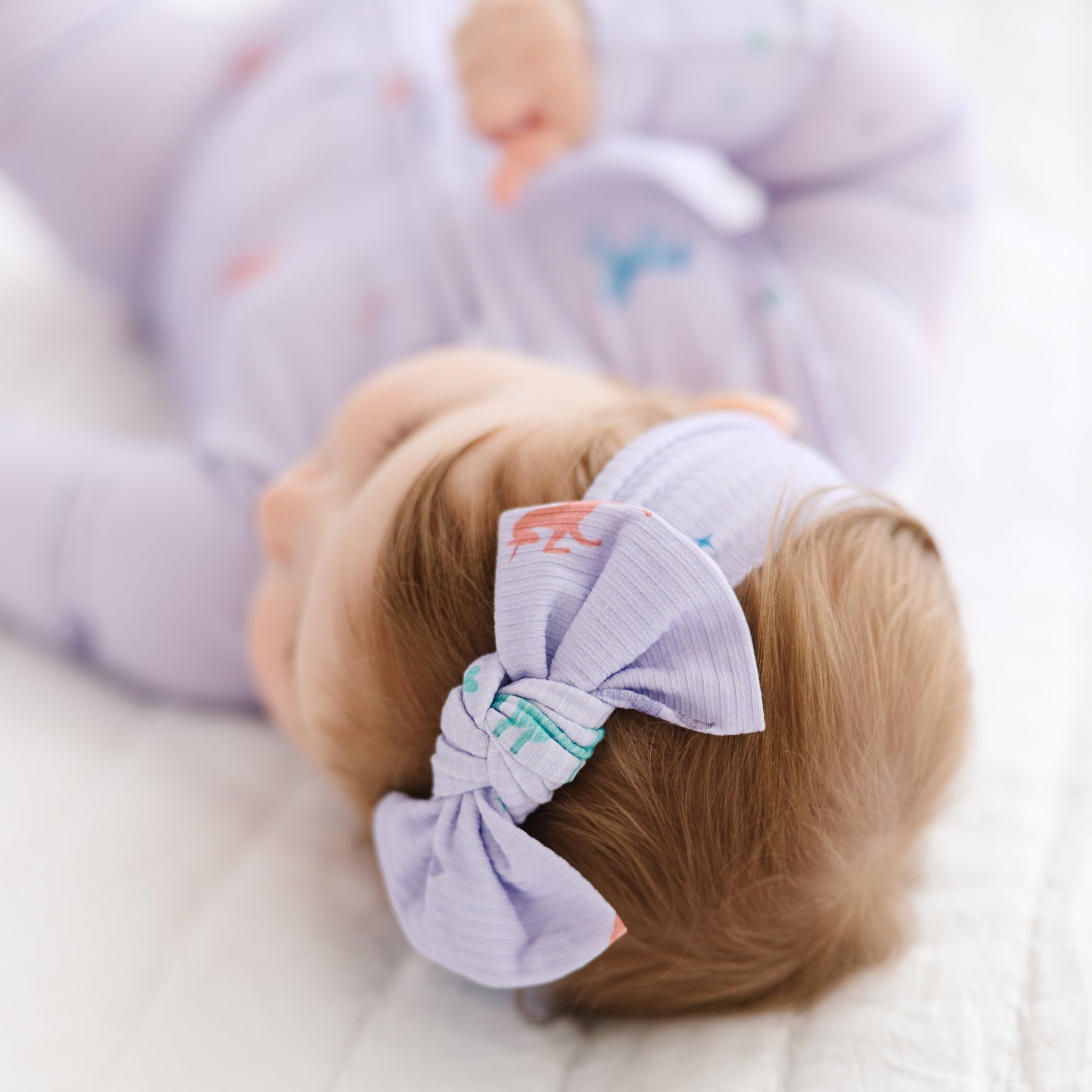 Baby with a purple bow in its hair, lying on a white blanket