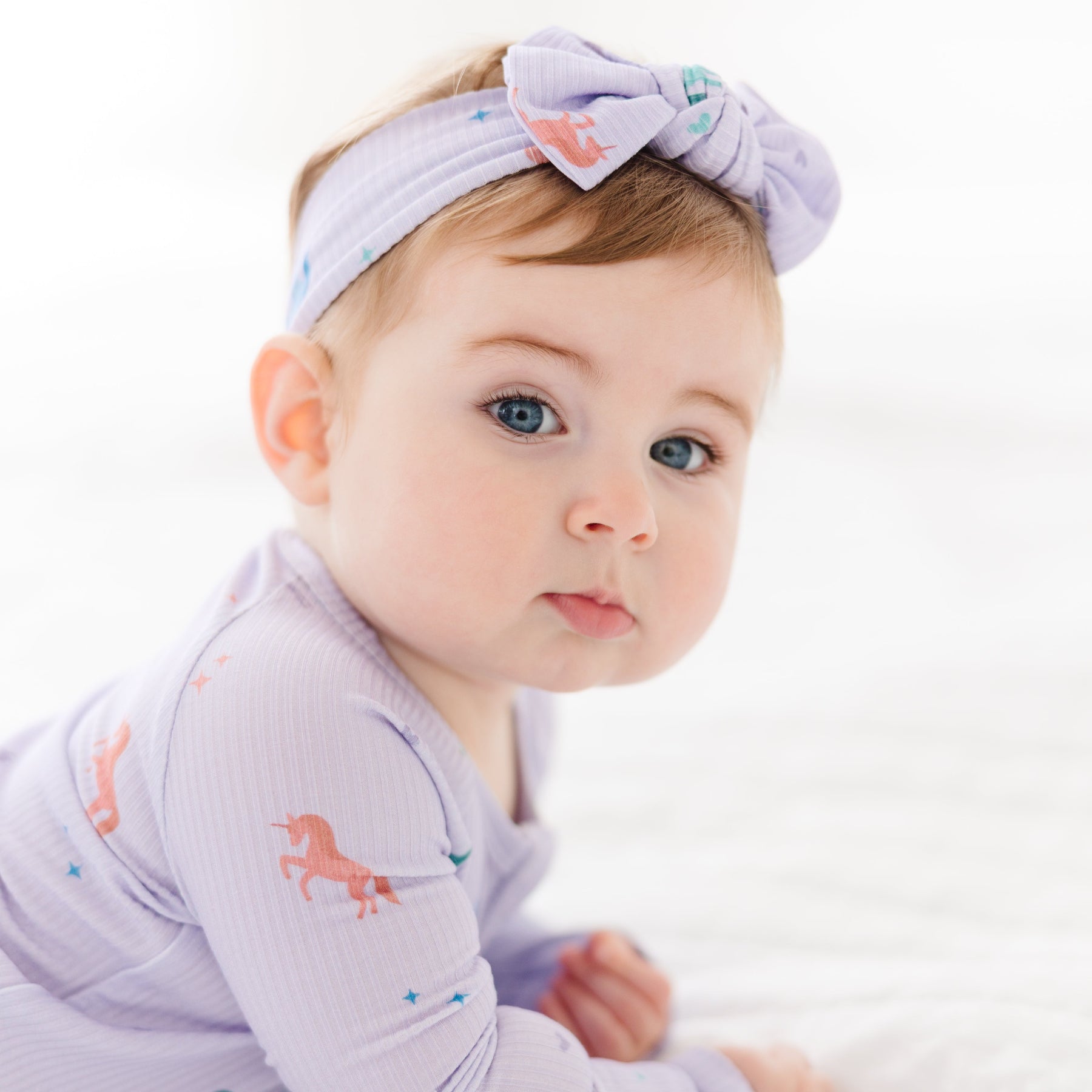 Baby wearing a light purple outfit with horse designs and a matching headband on a white background