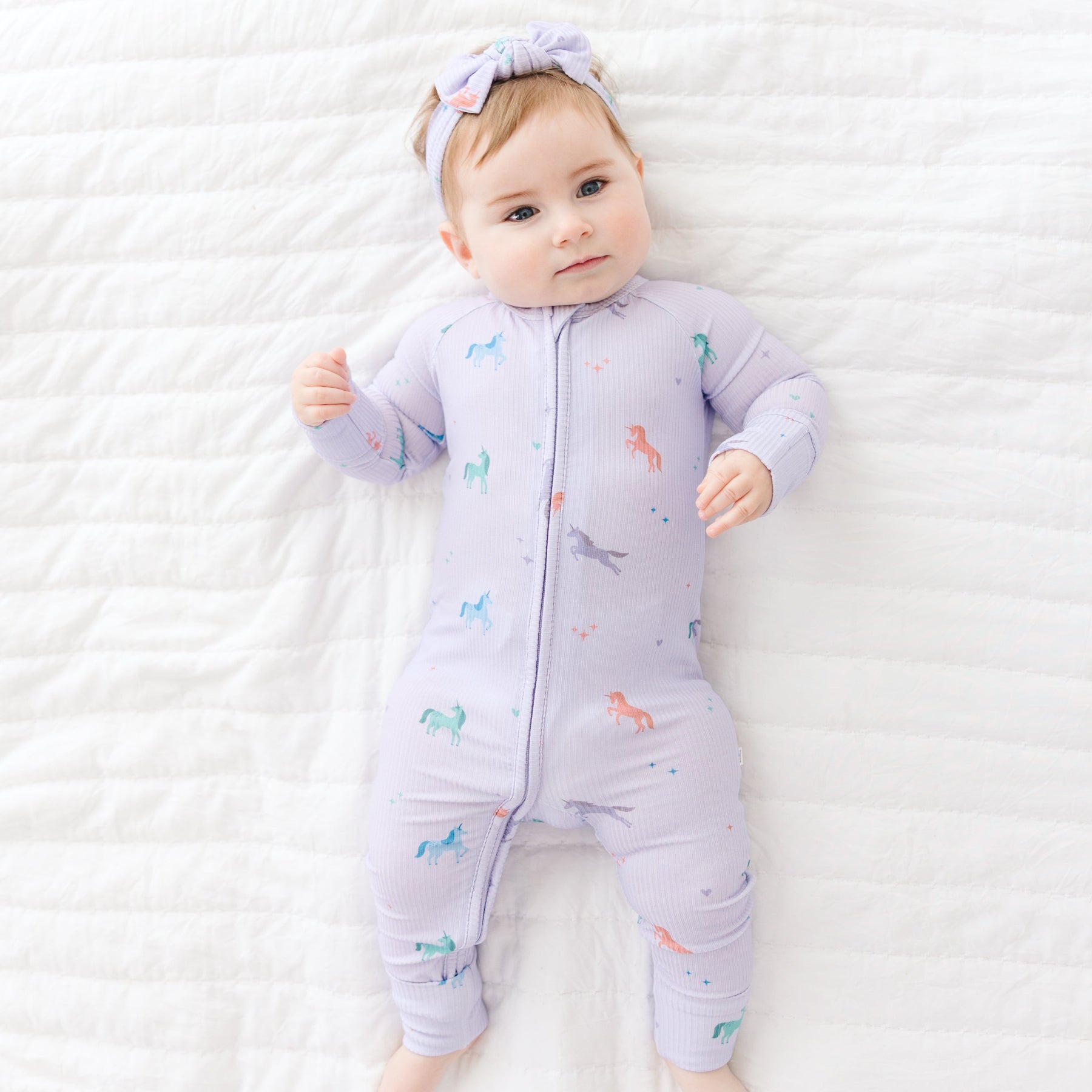 Baby wearing a purple Zip Romper with colorful unicorn designs on a white textured surface
