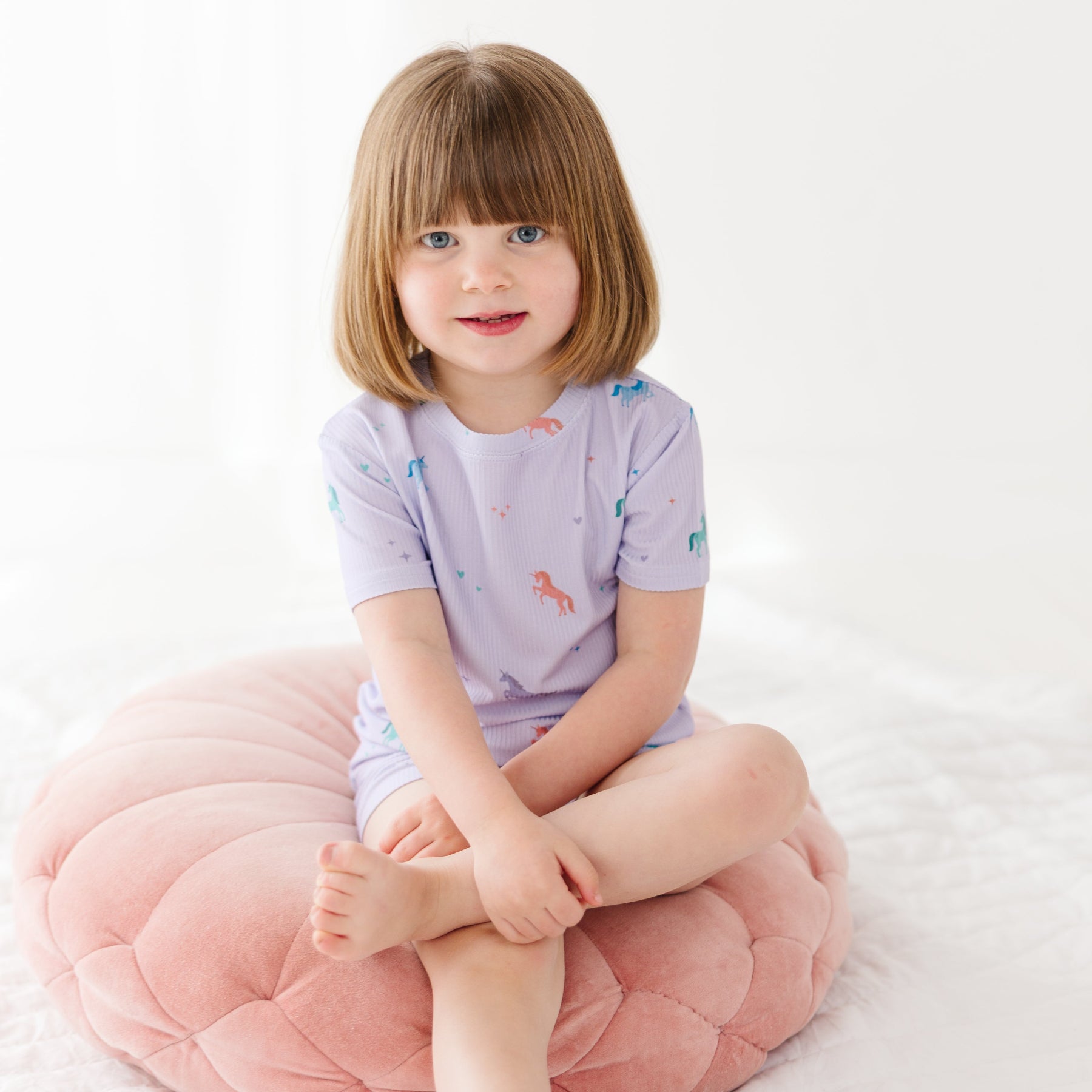 Young girl wearing a light purple pajama shorts set with unicorn patterns, sitting on a pink cushion with a white background