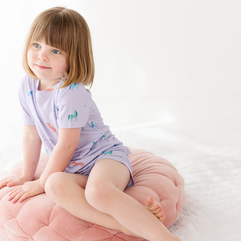Young girl wearing a light purple pajama shorts set with unicorn patterns, sitting on a pink cushion with a white background