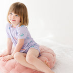Young girl wearing a light purple pajama shorts set with unicorn patterns, sitting on a pink cushion with a white background
