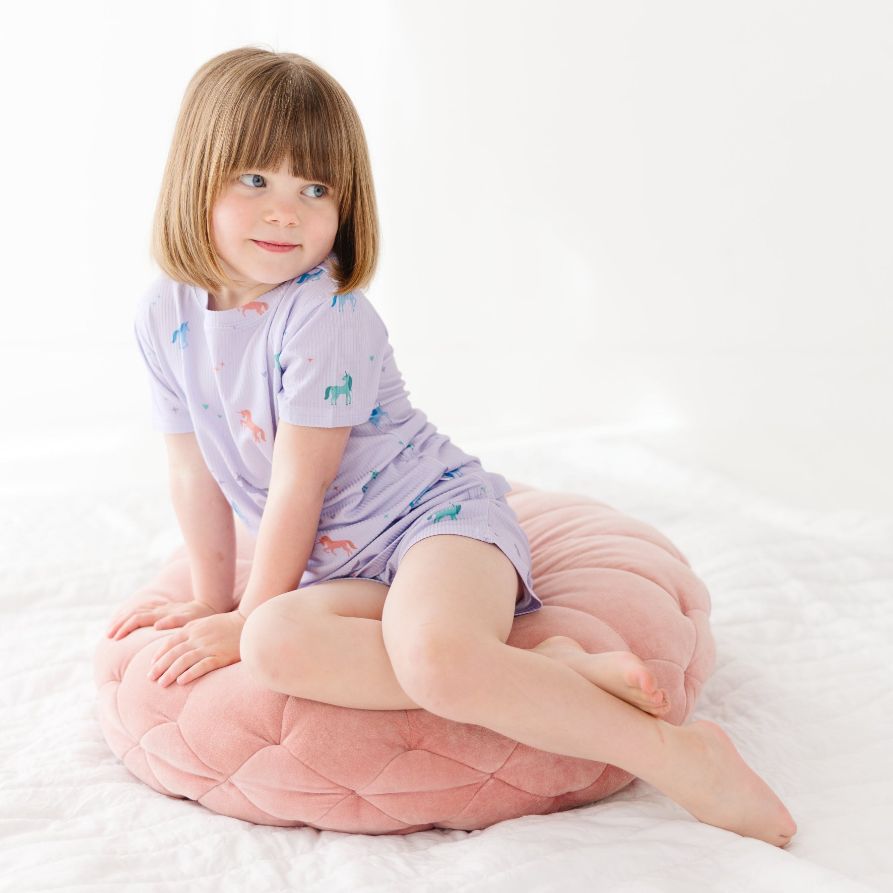 Young girl wearing a light purple pajama shorts set with unicorn patterns, sitting on a pink cushion with a white background