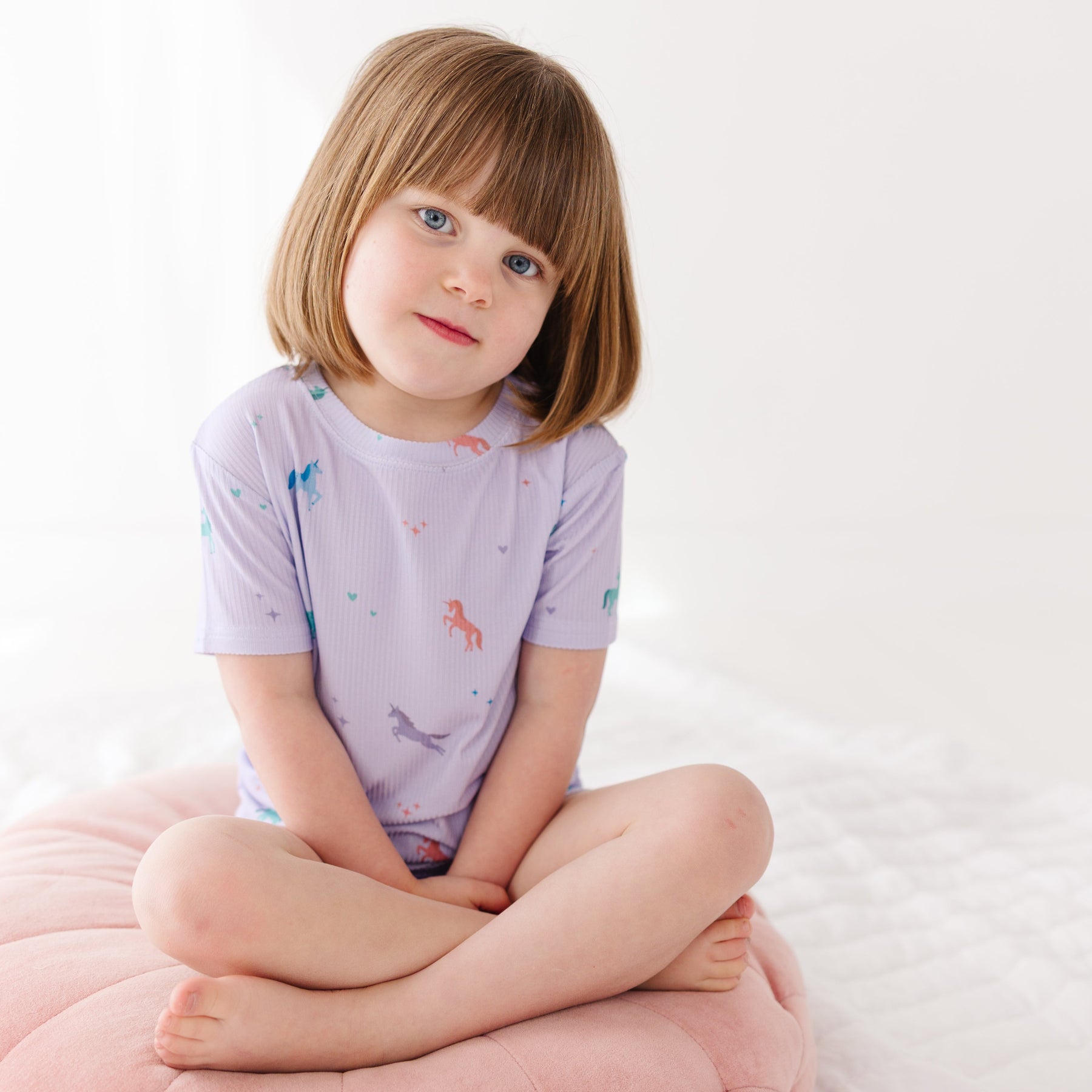 Young girl wearing a light purple pajama shorts set with unicorn patterns, sitting on a pink cushion with a white background