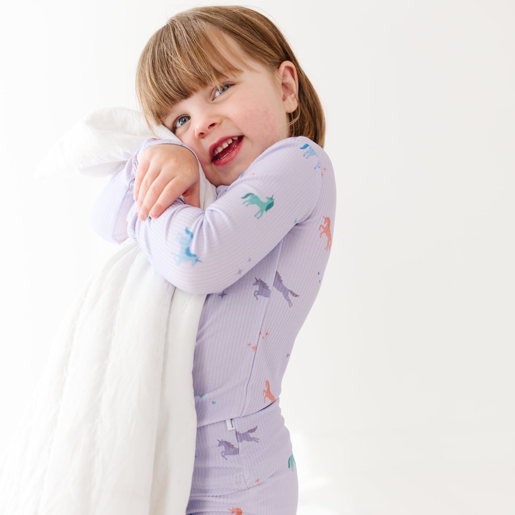 Child wearing a purple pajama set with unicorn prints, hugging a blanket, on a white background