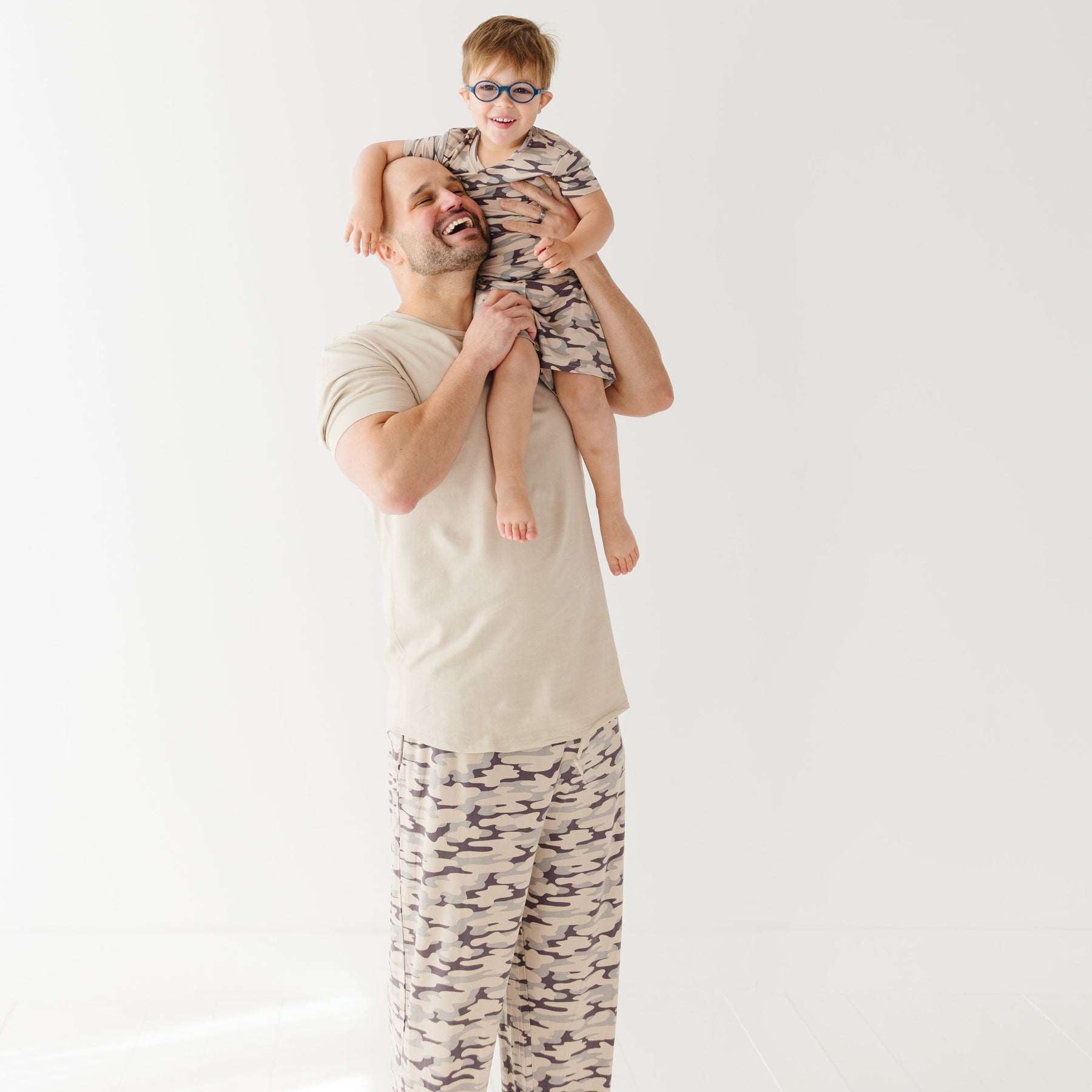 Man holding a child on his shoulders with matching camouflage outfits against a white background