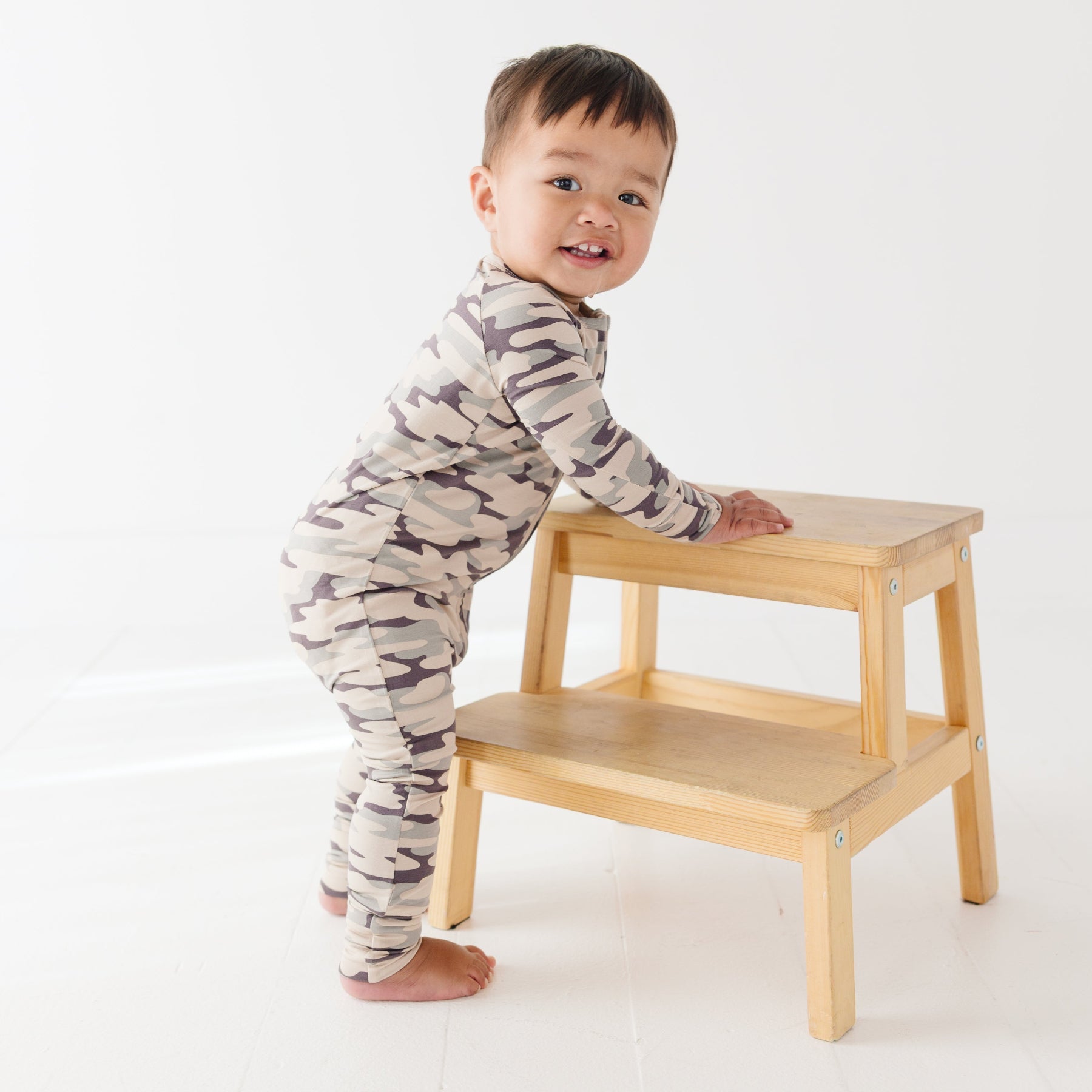 Baby wearing a camouflage onesie standing next on a wooden stool, on a white background