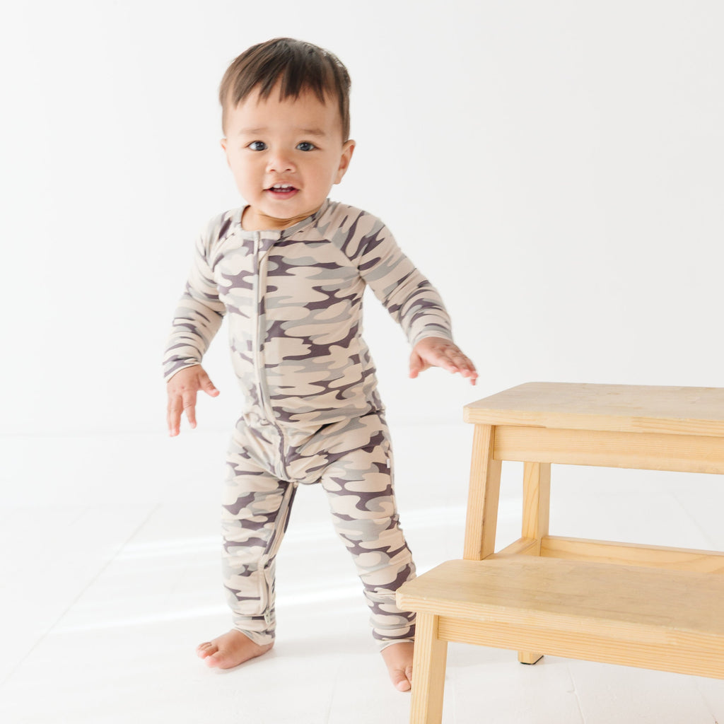 Baby wearing a camouflage onesie standing next on a wooden stool, on a white background