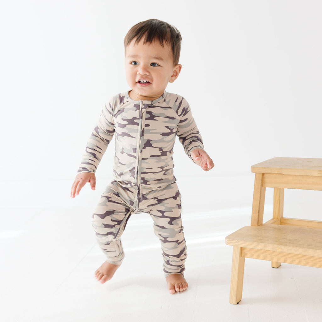 Baby wearing a camouflage onesie standing next on a wooden stool, on a white background