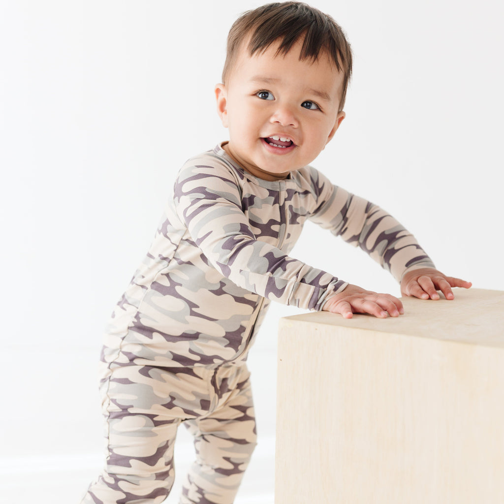 Baby wearing a camouflage onesie standing next on a wooden surface, on a white background