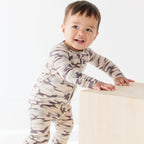 Baby wearing a camouflage onesie standing next on a wooden surface, on a white background