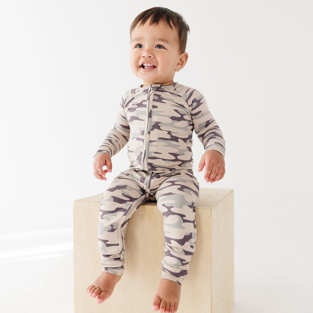 Baby wearing a camouflage onesie sitting on a wooden surface, on a white background