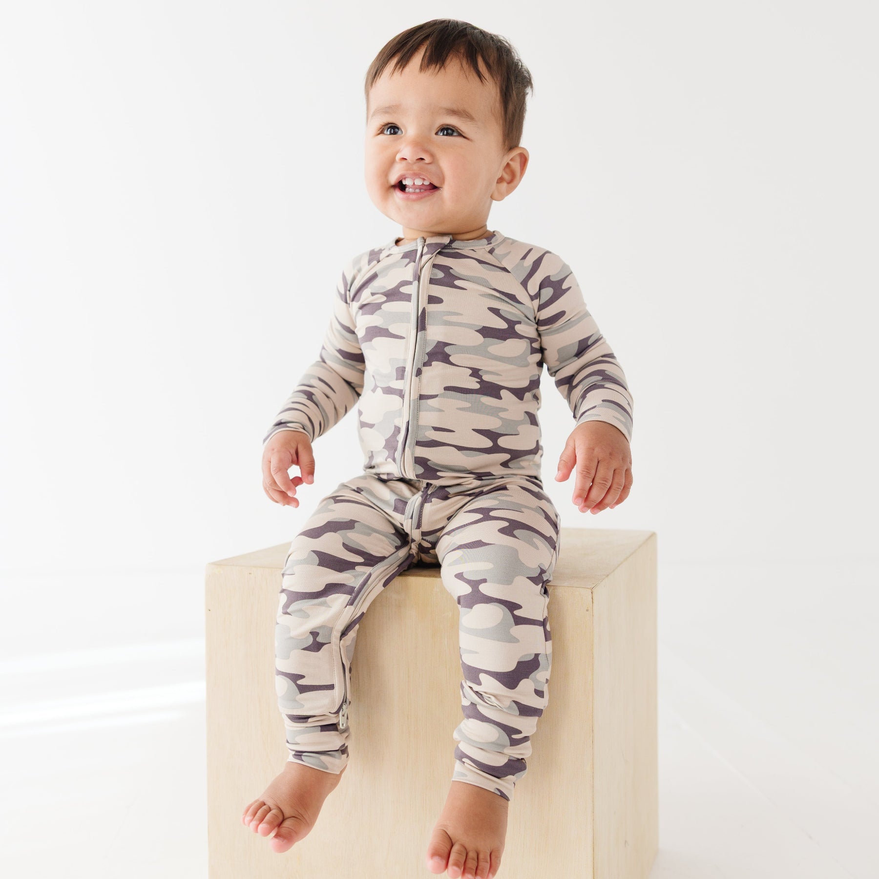 Baby wearing a camouflage onesie sitting on a wooden surface, on a white background