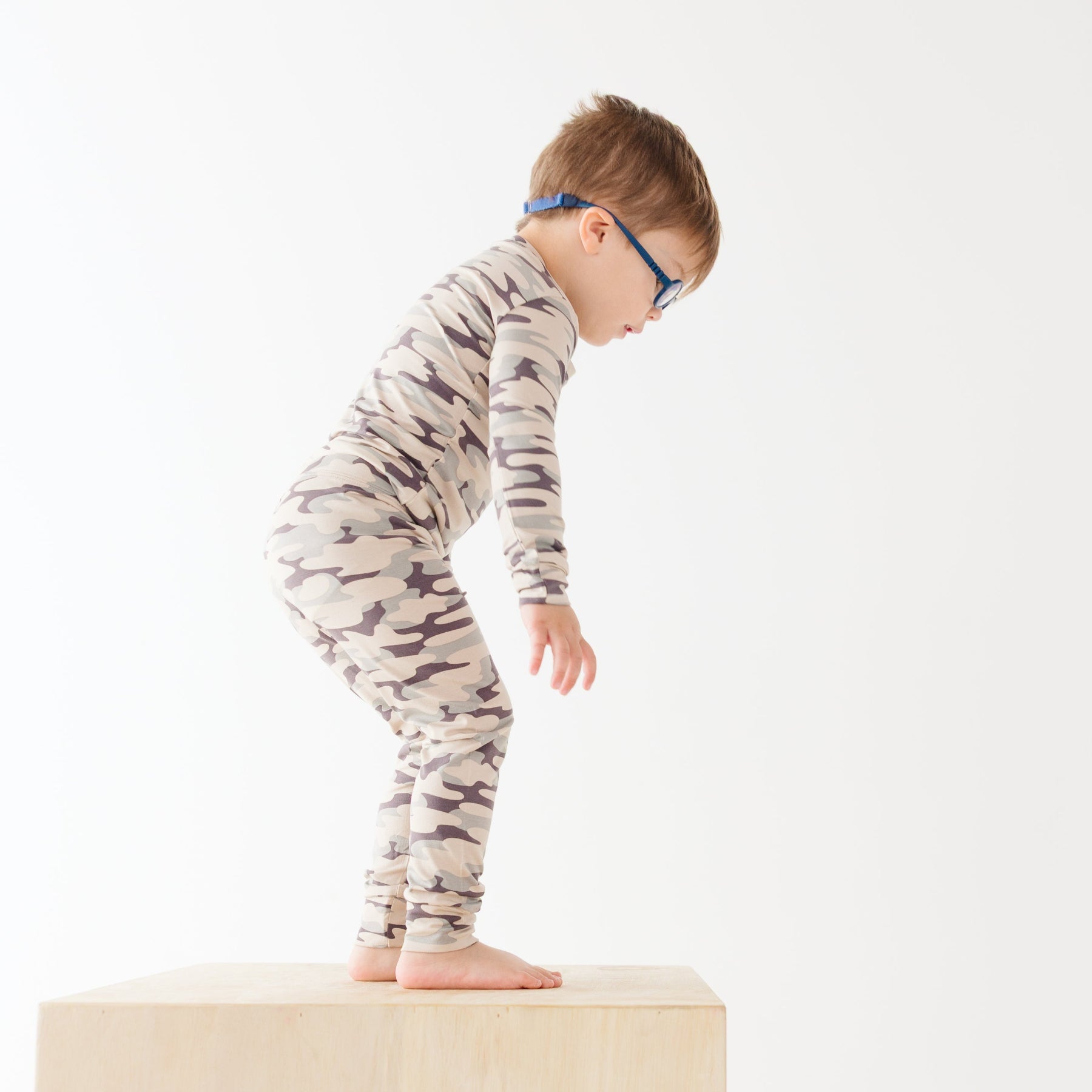 Child wearing a camouflage long sleeve pajama set standing on a wooden box on a white background