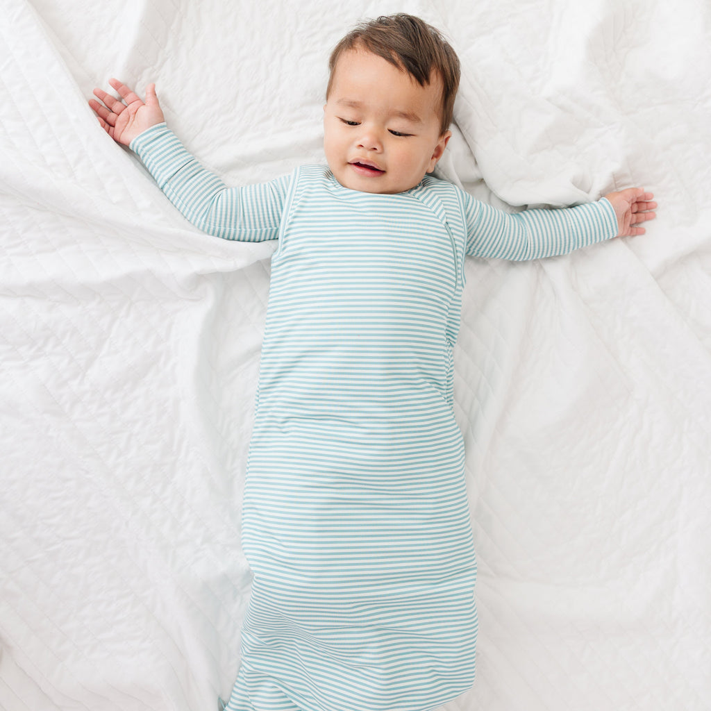 Baby lying on a white blanket wearing a light blue striped onesie and sleep sack.