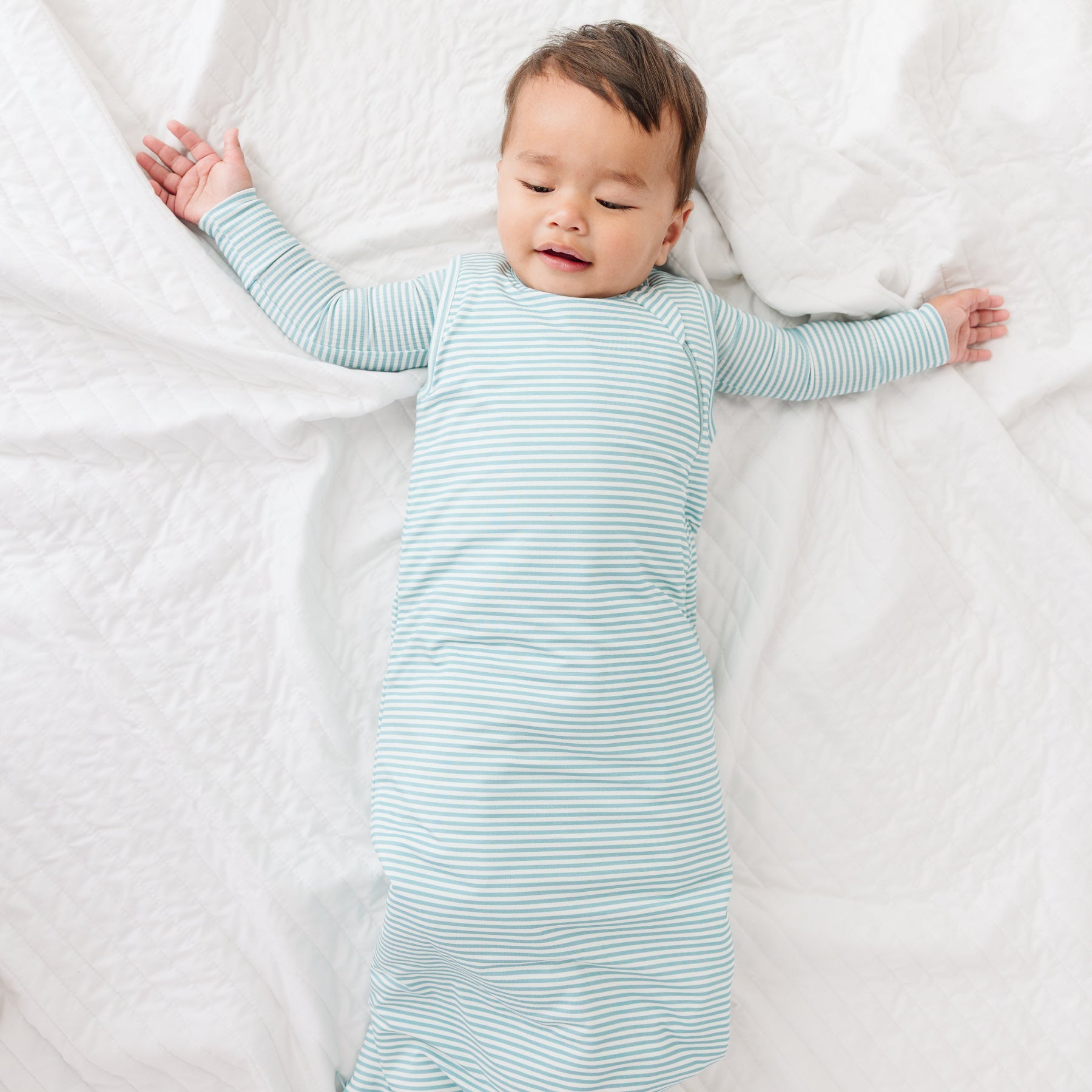 Baby lying on a white blanket wearing a light blue striped onesie and sleep sack.