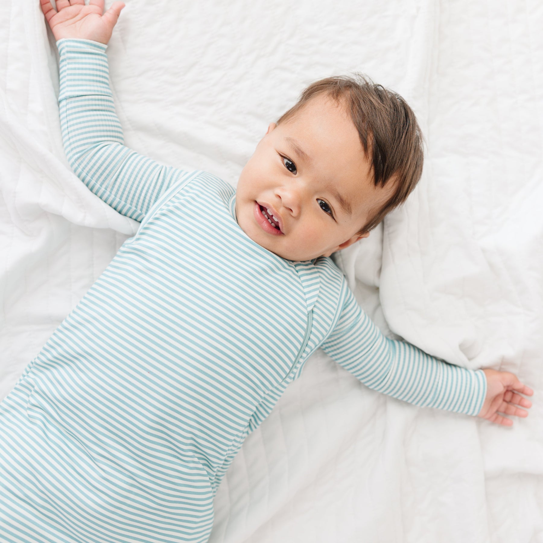 Baby lying on a white blanket wearing a light blue striped onesie and sleep sack.