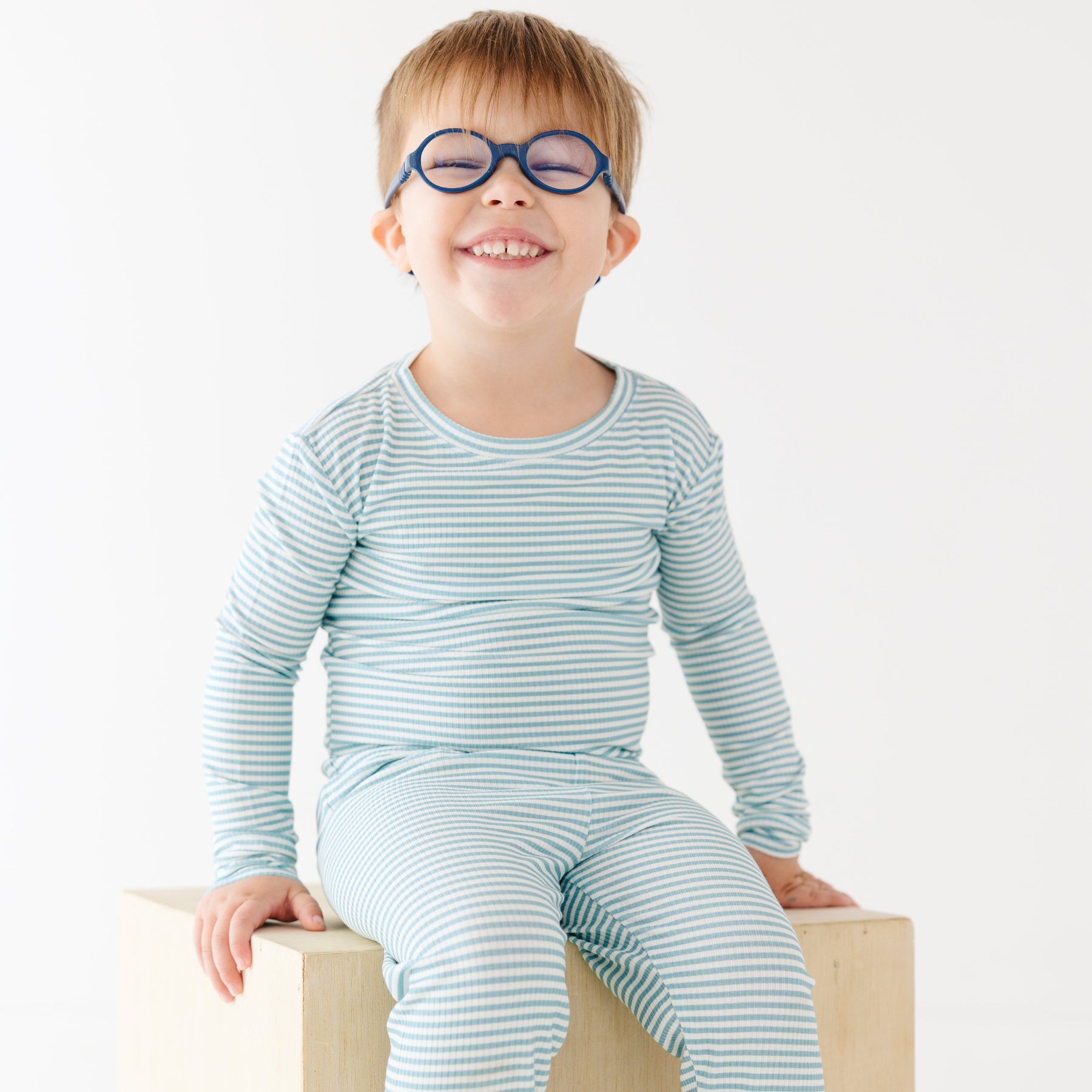 Child wearing a light blue and white striped outfit sitting on a wooden block against a white background