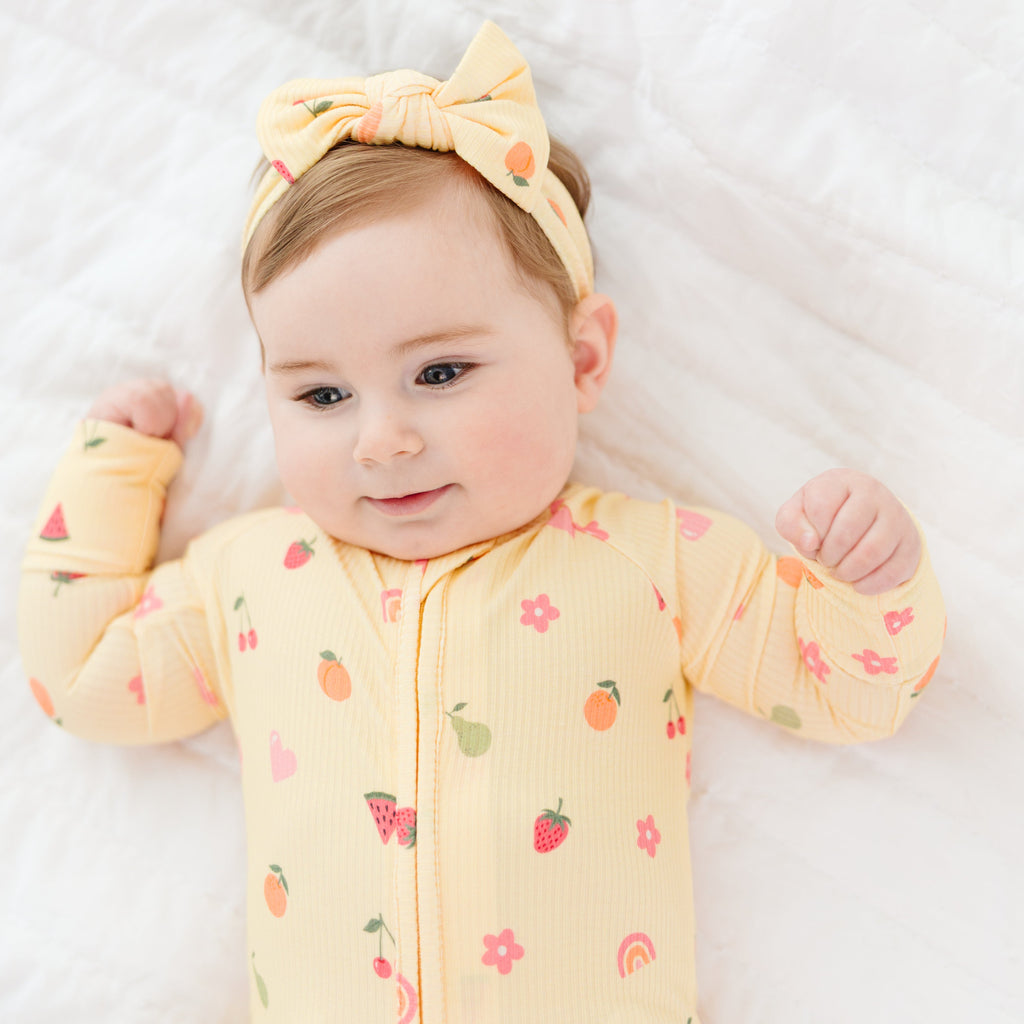 Baby wearing a yellow outfit with fruits patterns and a matching headband on a white background