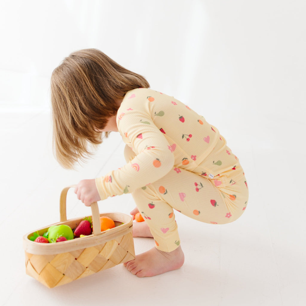 Child wearing a yellow pajama set with heart patterns playing with a basket of fruits on a white background