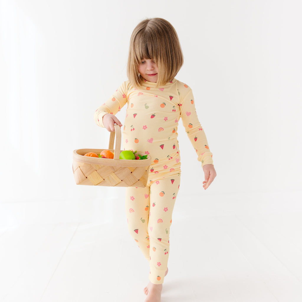 Child wearing a yellow pajama set with fruit pattern, holding a basket of fruits on a white background