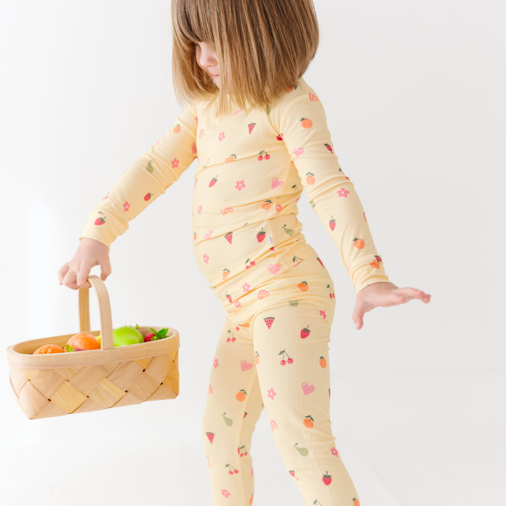 Child wearing a yellow pajama set with fruit pattern, holding a basket of fruits on a white background