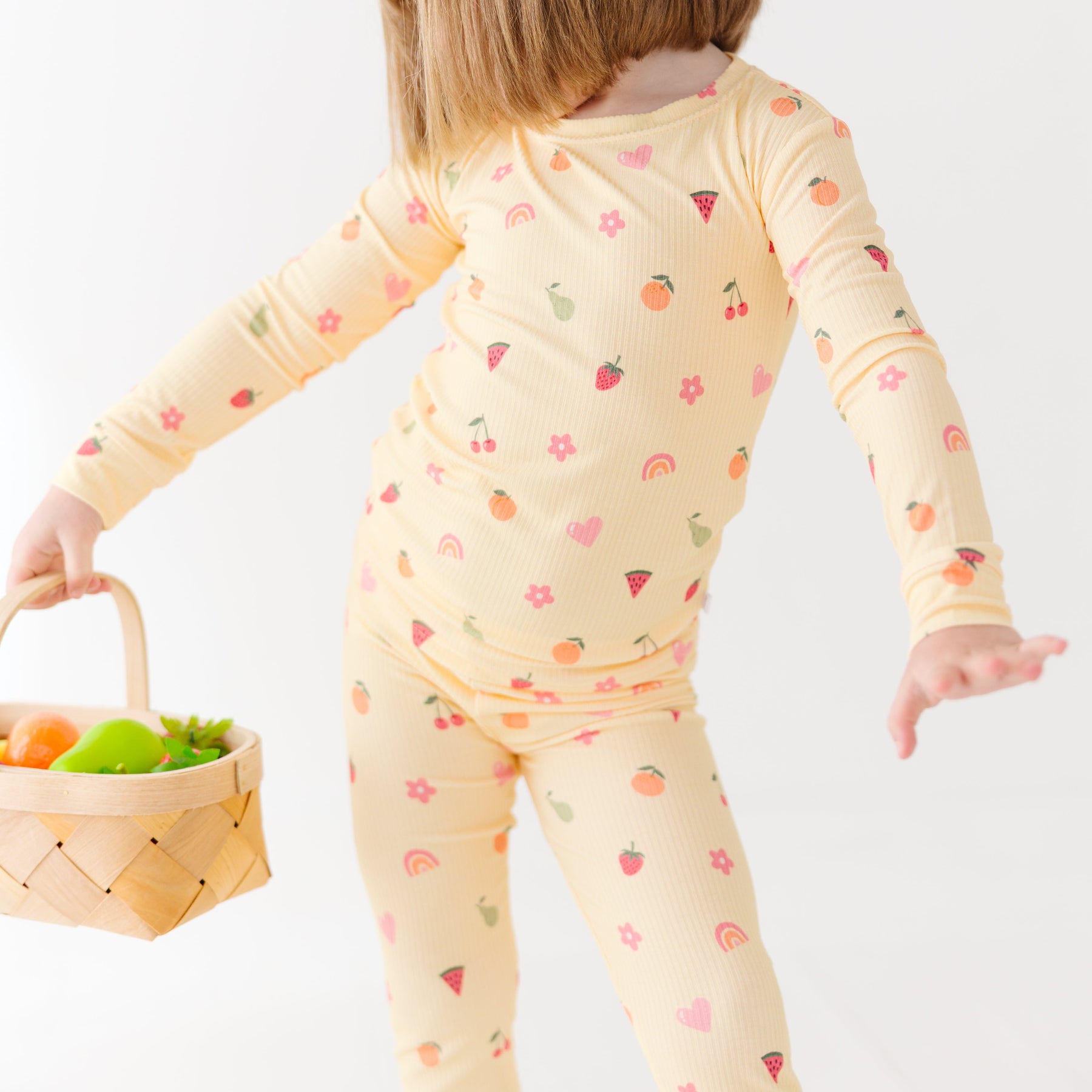Child wearing a yellow pajama set with fruit patterns, holding a basket of fruits on a white background