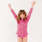 Young girl wearing a red and white floral swimsuit with arms raised on a white background