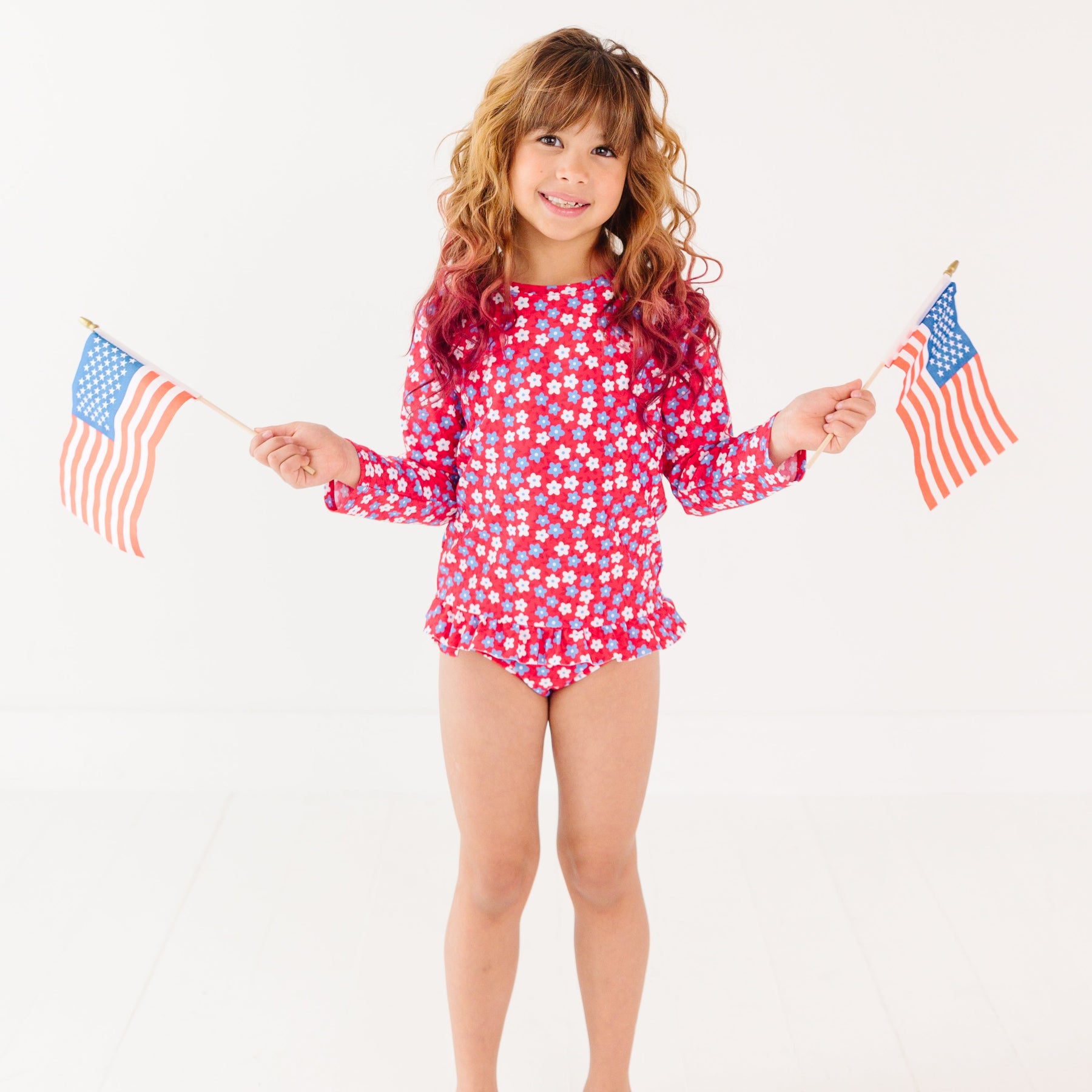 Young girl wearing a red and white floral swimsuit holding two small American flags on a white background