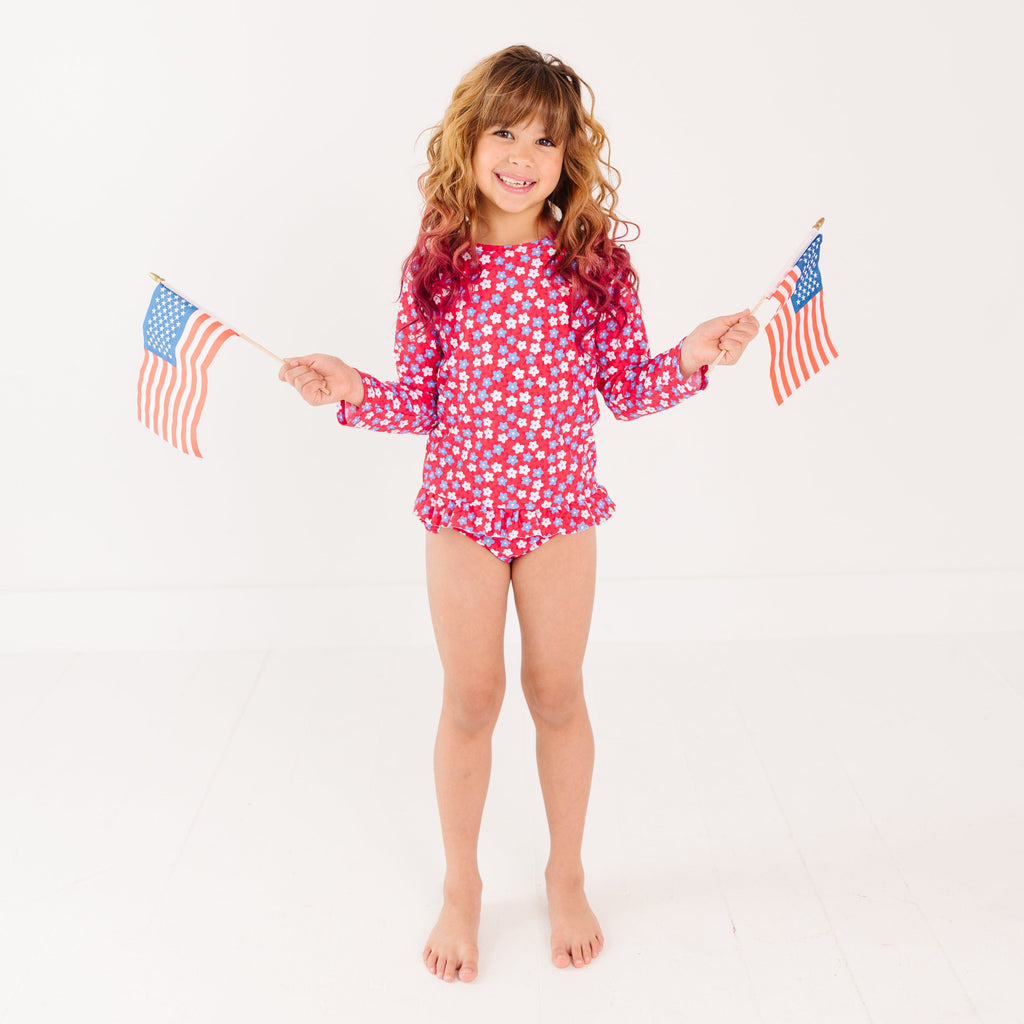 Young girl wearing a red and white floral swimsuit holding two small American flags on a white background