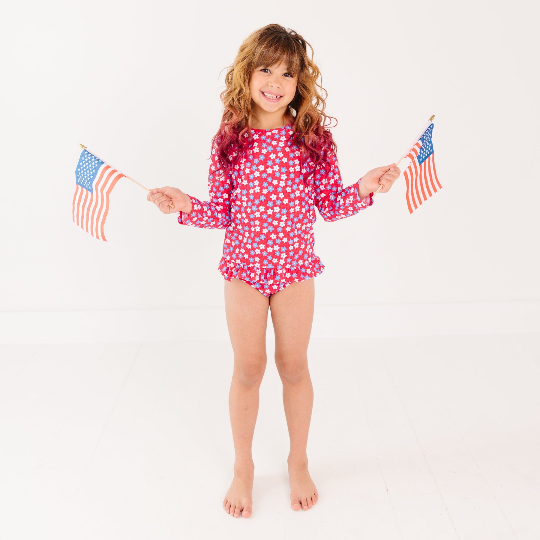 Young girl wearing a red and white floral swimsuit holding two small American flags on a white background