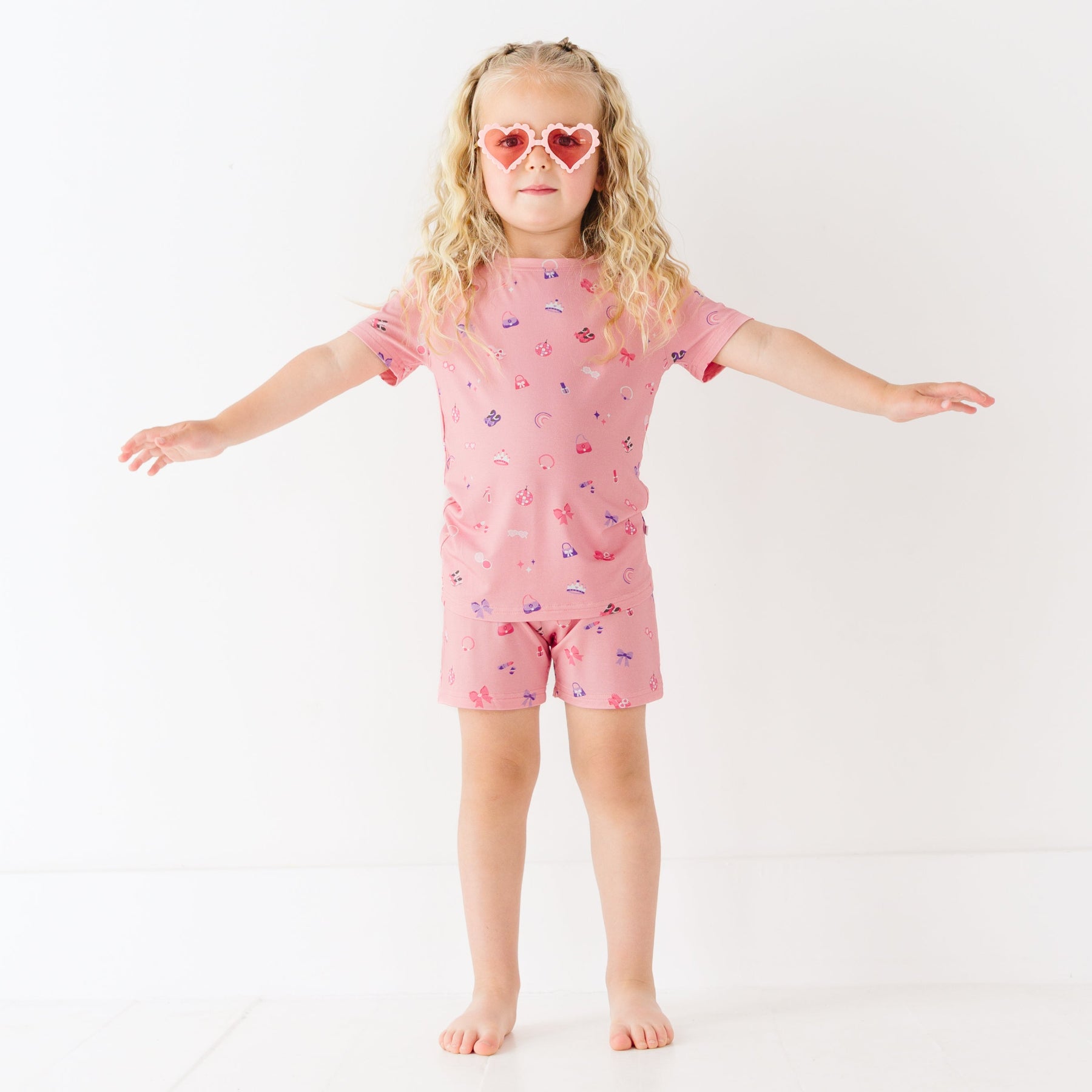 Child wearing a pink shorts pajama set with heart sunglasses on a white background