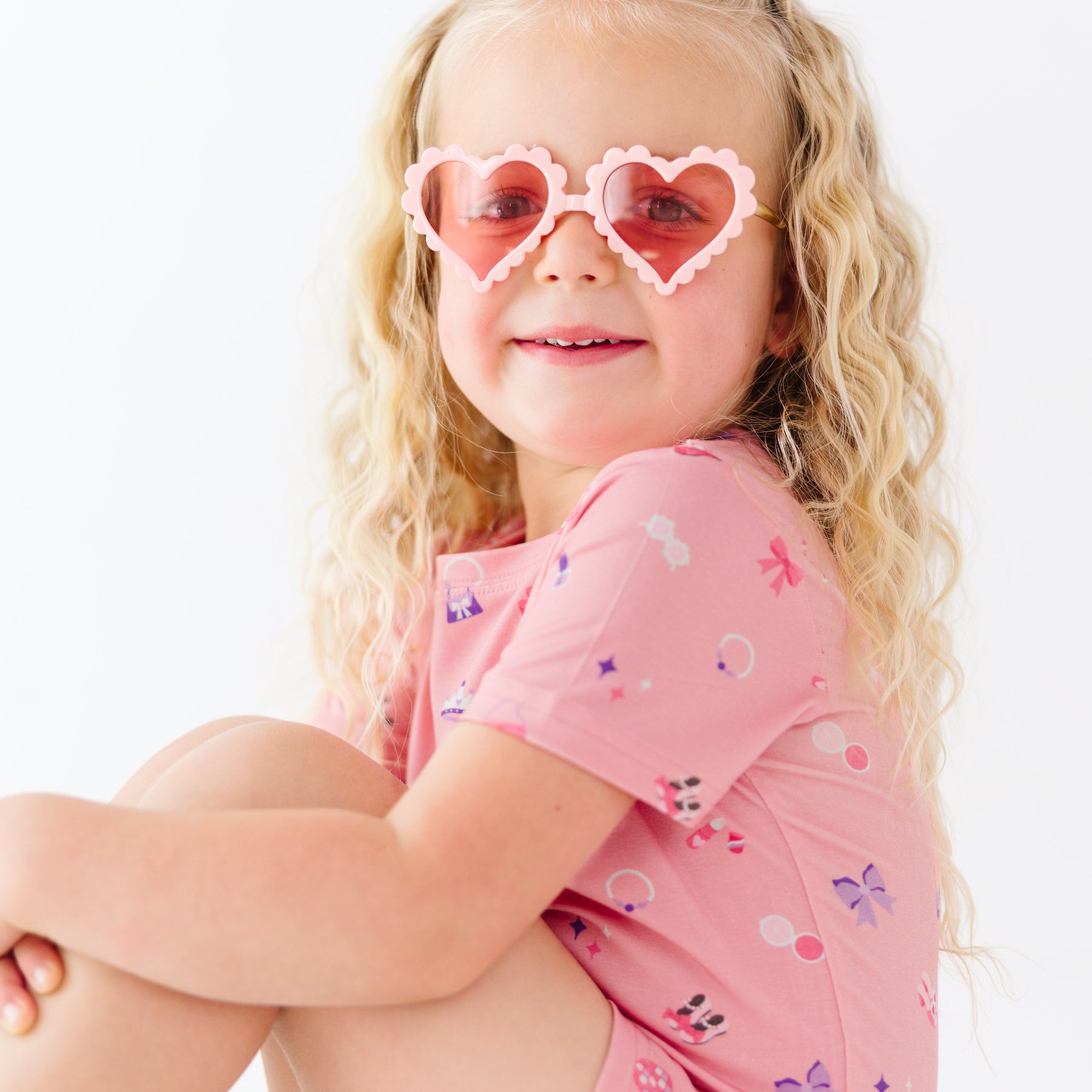 Child wearing a pink outfit with girly patterns and heart-shaped sunglasses on a white background