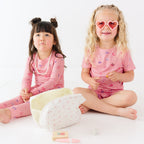 Two children in pink pajamas sitting on a white surface with a small bag beside them.