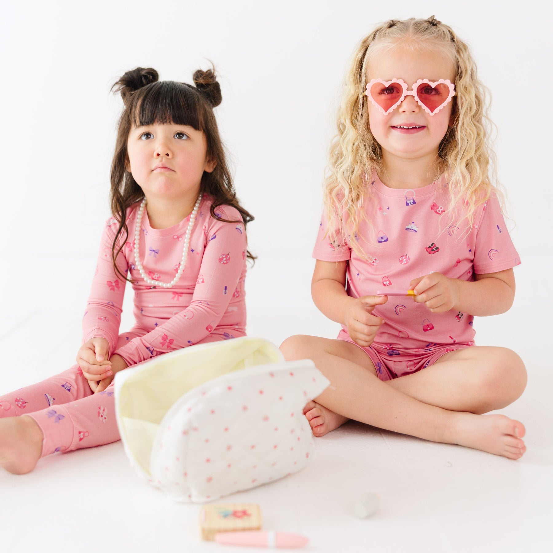 Two children in pink pajamas sitting on a white surface with a small bag beside them.
