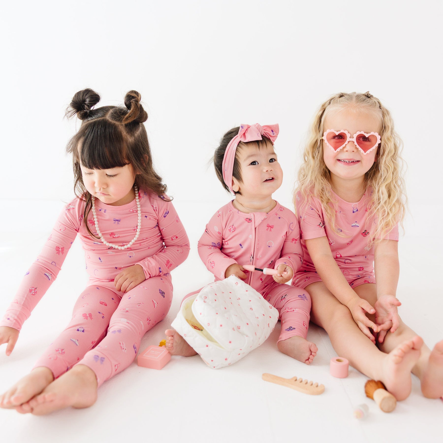Three children in pink pajamas sitting on a white surface with toys around them.