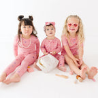 Three children in pink outfits sitting on a white surface with a white background
