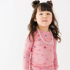 Young girl wearing a pink long-sleeve shirt with girly patterns and a pearl necklace on a white background