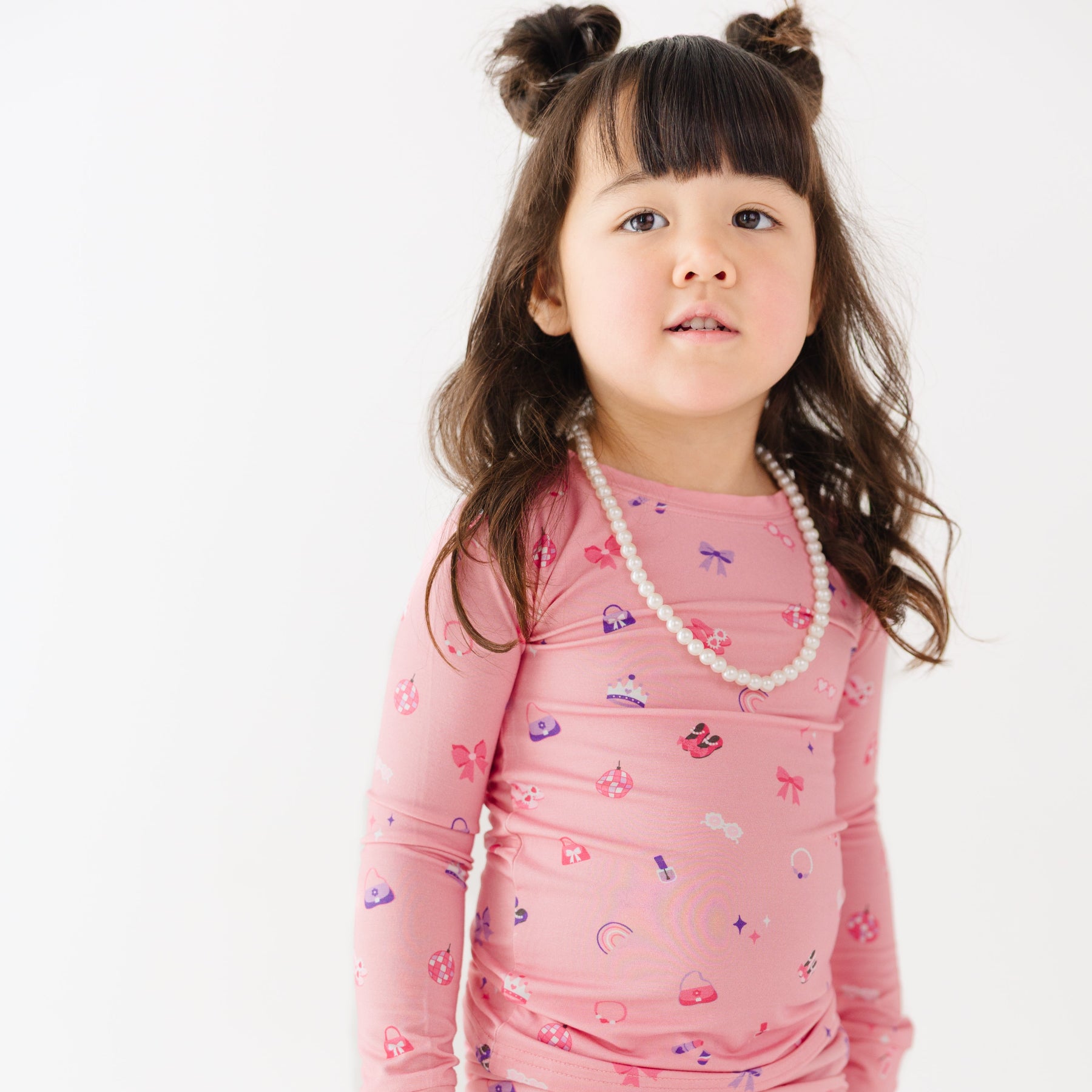 Young girl wearing a pink long-sleeve shirt with girly patterns and a pearl necklace on a white background