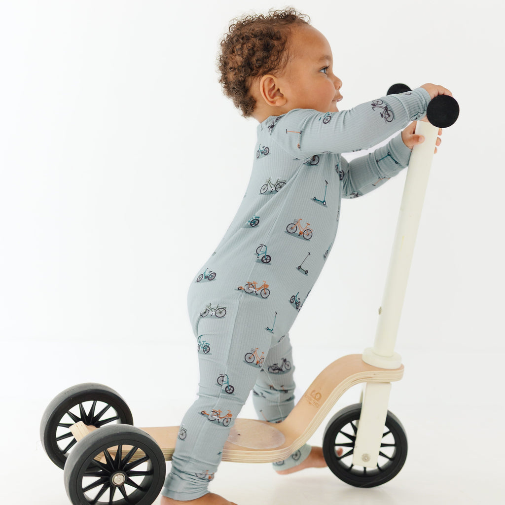 Child wearing a light blue onesie with vehicle patterns, standing on a wooden scooter with large wheels.