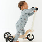 Child wearing a light blue onesie with vehicle patterns, standing on a wooden scooter with large wheels.