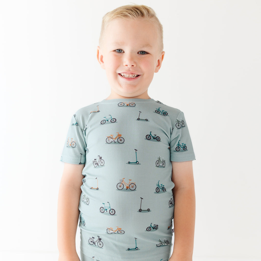Child wearing a light blue outfit with bicycles & scooter patterns on a plain background