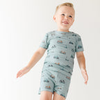 Child wearing a light blue outfit with bicycles & scooter patterns on a plain background