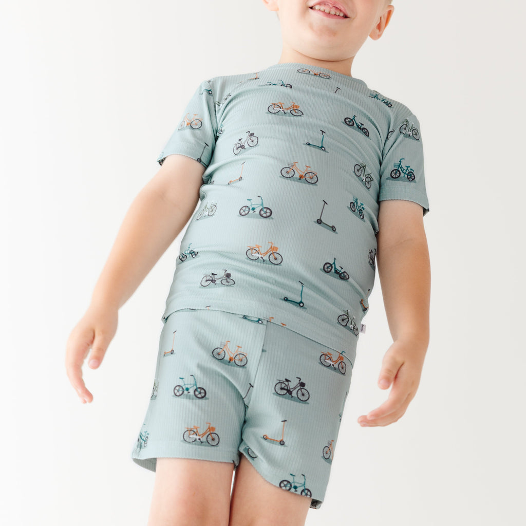 Child wearing a light blue outfit with bicycles & scooter patterns on a plain background