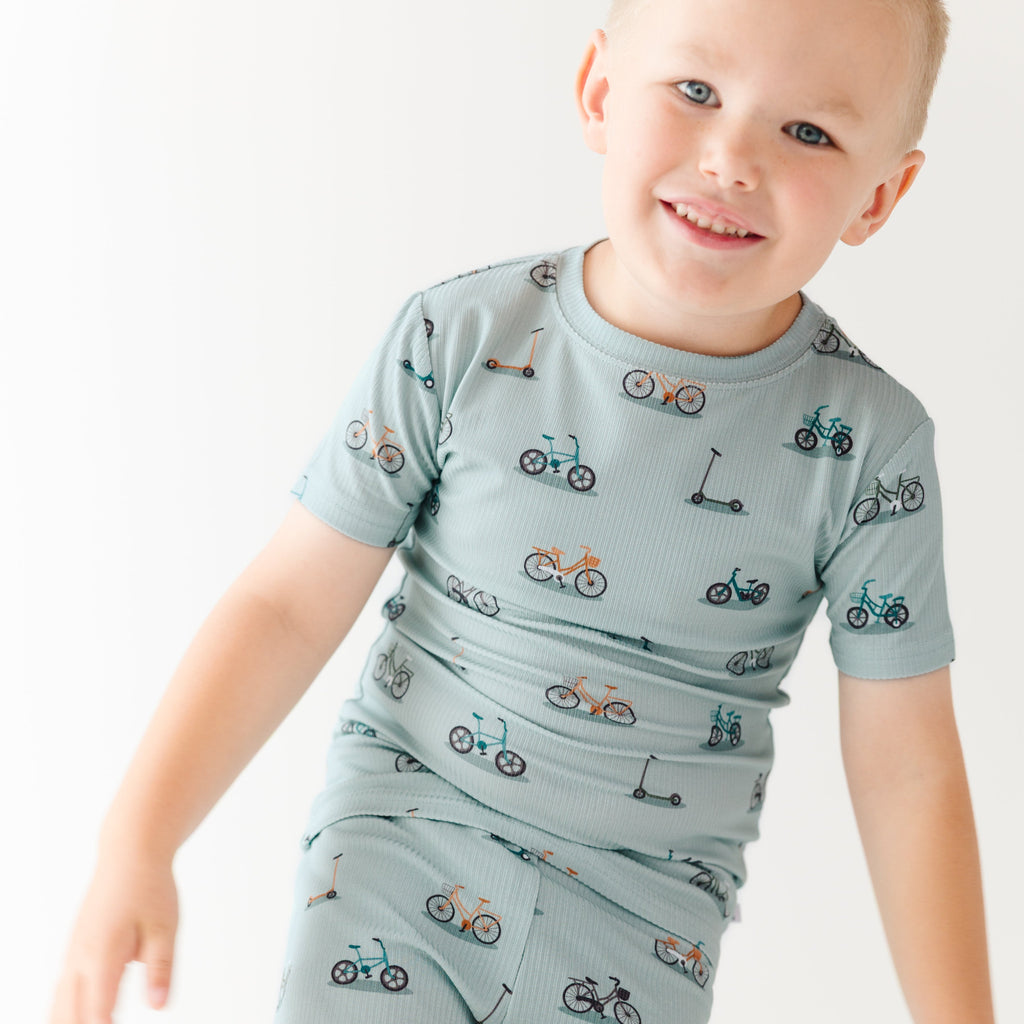 Child wearing a light blue outfit with bicycles & scooter patterns on a plain background