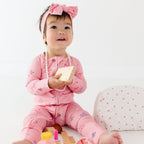 Baby in pink onesie with bow headband sitting on a white surface