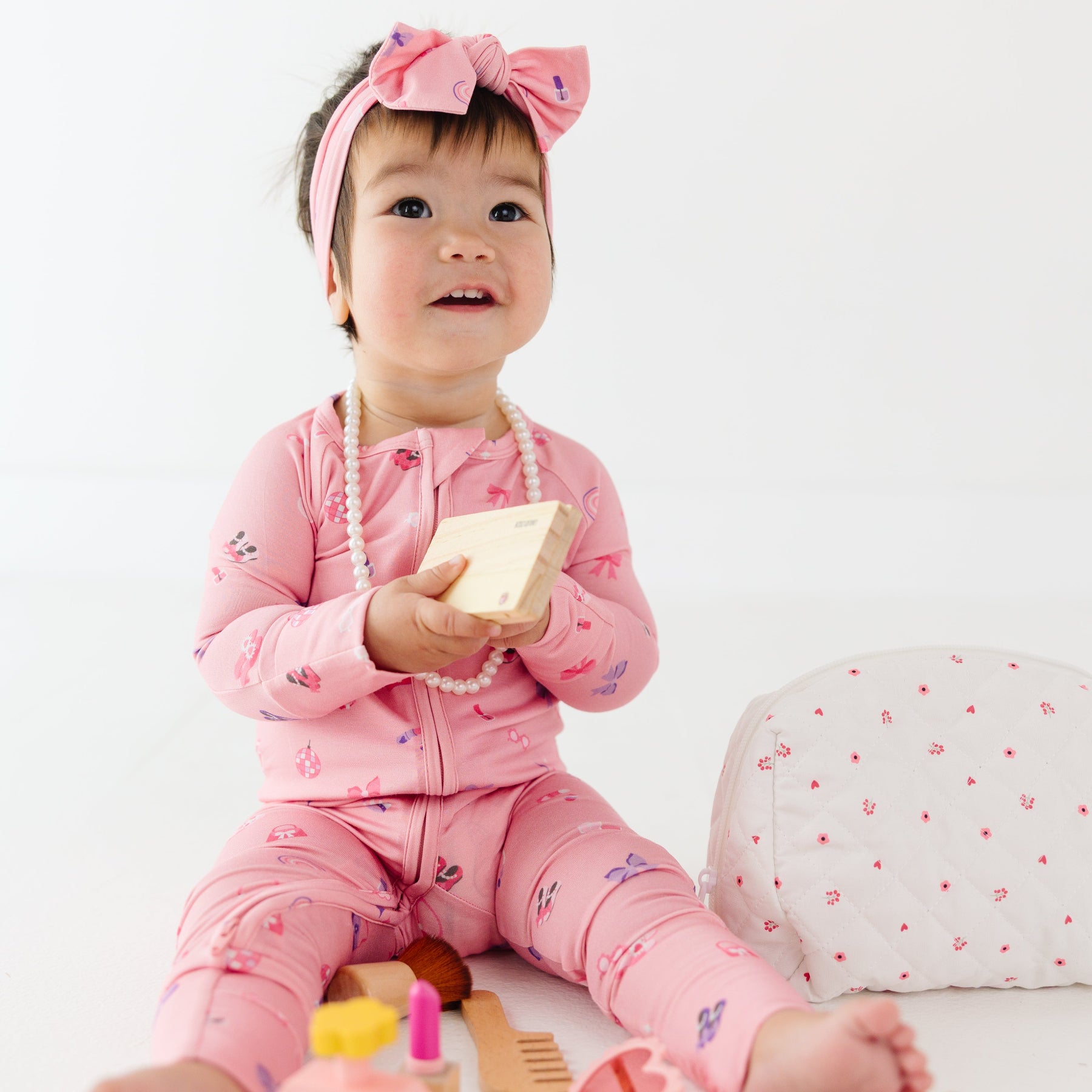 Baby in pink onesie with bow headband sitting on a white surface