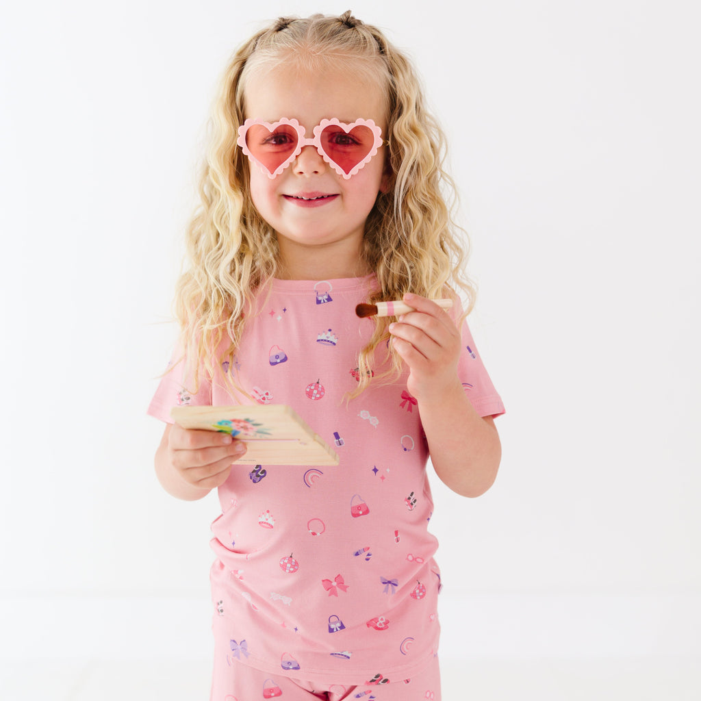 Young girl wearing pink sunglasses with heart shapes, holding a brush and make-up palette on a white background
