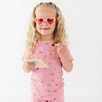 Young girl wearing pink sunglasses with heart shapes, holding a brush and make-up palette on a white background